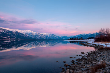 Fototapeta premium A serene waterscape, the glassy surface reflecting snow-capped peaks and pastel skies at dawn's first light. Shoreline in foreground