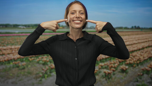 Woman points finger to cheeks in a tulip field outdoors near rows of flowers under blue sky; playful joy.