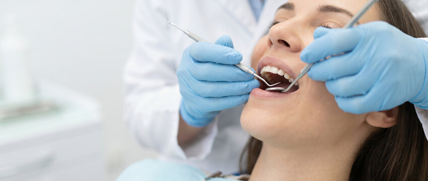 uma jovem mulher brasileira recebendo tratamento odontol&oacute;gico em cl&iacute;nica moderna. M&atilde;os de dentista com luvas azuis examinando os dentes da paciente. Imagem com espa&ccedil;o para c&oacute;pia, ideal para banner 