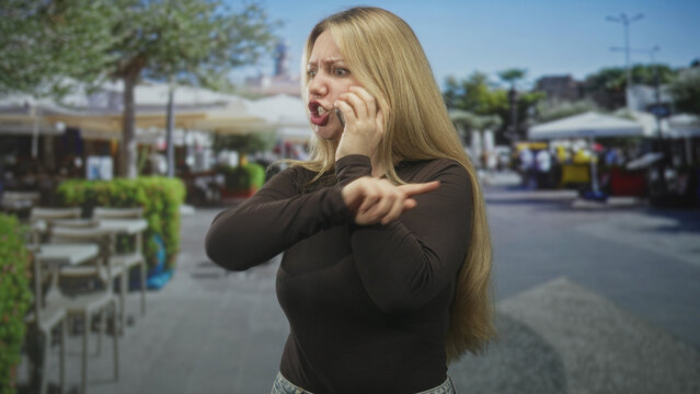 Woman blonde russian caucasian talking on phone, pointing finger and clenching fist with scowl in a busy street cafe by outdoor tables and umbrellas; anger conflict.