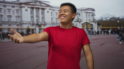 Fototapeta premium Man in red t shirt with outstretched hand and visible forearm on busy street in front of ornate building smiling toward camera; welcoming joy.