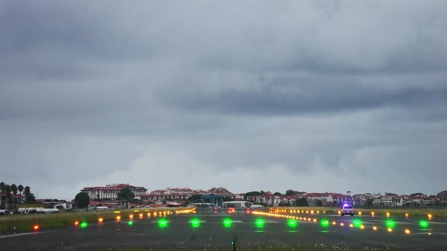 Wide shot of an airport runway threshold at dusk showing rows of green, amber, and red runway lights stretching into the distance under a dramatic stormy sky, with an emergency vehicle's blue flashing