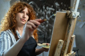 Fotobehang Schilderij Curly woman painter using spatula while painting adding a touch to her artwork working in creative art studio  © Andrii Lysenko