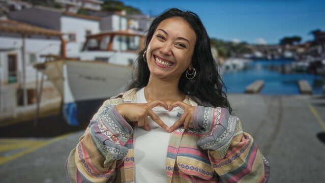 Woman points finger to camera and forms heart with hands on a seaside street beside moored boats and dock buildings; joy affection.