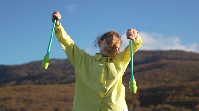 Gymnastics girl clubs practicing rhythmic performance on windy mountain hilltop against clear blue sky background.