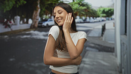 Obraz premium Young hispanic woman in white crop top, eyes closed, hand resting on cheek and arms crossed on a tree lined urban street near parked cars; contentment self care.