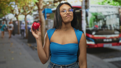 Fototapeta premium Woman holding red heart prop on a busy city street beside a parked bus and pedestrians; health awareness playful.