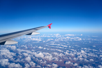 Fototapeta premium An airplane wing stretches into a brilliant blue sky, overlooking a wide expanse of white cumulus clouds scattered over the landscape below during daytime flight