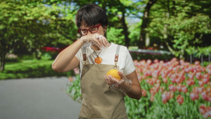Fototapeta premium Woman wearing apron holds an orange and wipes her nose with a gloved hand in studio; concern hygiene.