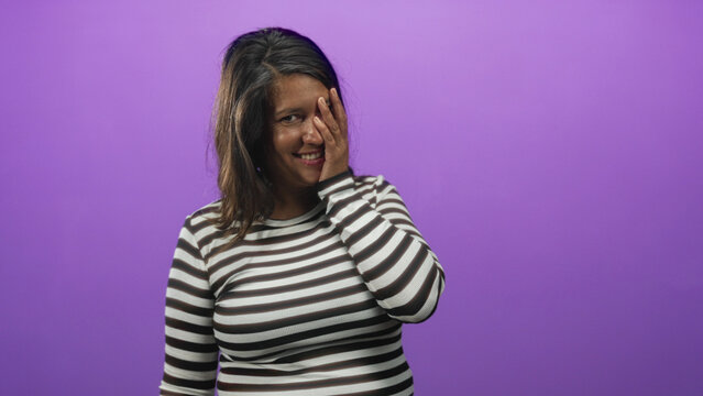 Woman covers face with hand while smiling and tilting her head in a purple studio with a seamless backdrop wearing a striped shirt; bashful joy.