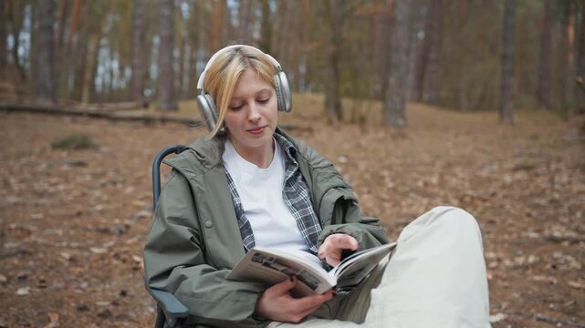 Blonde Caucasian woman reading book while wearing headphones in forest. Female traveler relaxing on folding chair during camping vacation. Peaceful weekend recreation outdoors in nature.