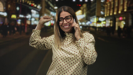 Fototapeta premium Woman holds phone to ear and raises arm in celebration on a busy city street at night; joy celebration success.