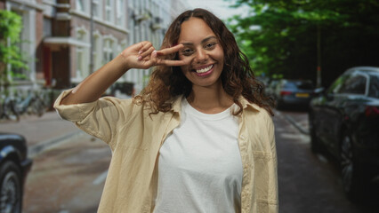 Fototapeta premium Woman making a peace sign with fingers at her eye while smiling in an urban street scene with parked cars and bicycles visible; playful joy.