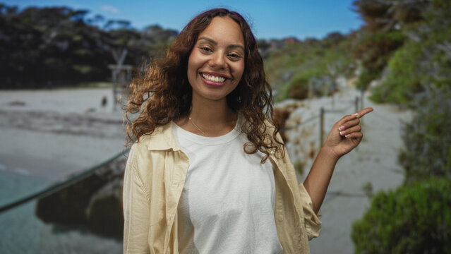 Young african american woman smiling, finger pointing to right on sandy seaside path with lifeguard tower visible; joy travel.