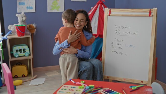 Teacher hugs little boy beside whiteboard and toys in a child learning corner inside a building; gentle comfort.