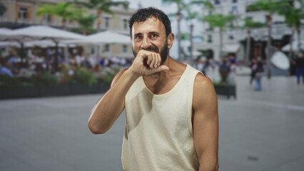 Fototapeta premium Man biting his fist, hand to mouth gesture in a busy street cafe wearing a sleeveless tanktop, bearded middleaged hispanic; stifled amusement.