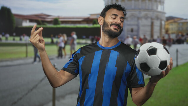 Man hispanic middle age with beard in blue striped soccer uniform holding a soccer ball in one hand and making a pinched fingers gesture at building lawn and walkway; confidence swagger.