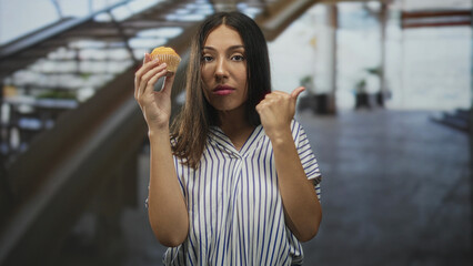 Fototapeta premium Hispanic brunette woman holding a muffin in one hand while thumb pointing to the side with the other, standing near an escalator in a building; skepticism reluctance.