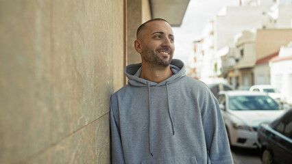 Obraz premium Man leans against stone wall smiling, wearing gray hoodie and earring, casual pose on a sunny street; serenity.