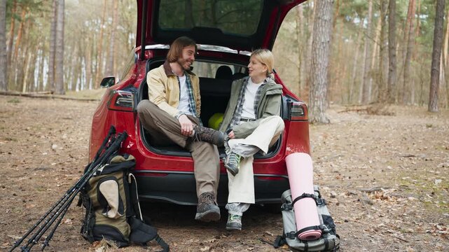 Caucasian couple talking with each other while sitting inside open car trunk in forest. Blonde woman and handsome man resting near hiking backpacks. Travelers enjoying camping weekend in nature.