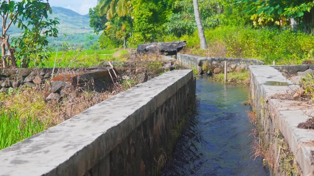 Close up shot of clear water flowing in a concrete irrigation ditch surrounded by green tropical trees and hills in rural Flores, Indonesia.
