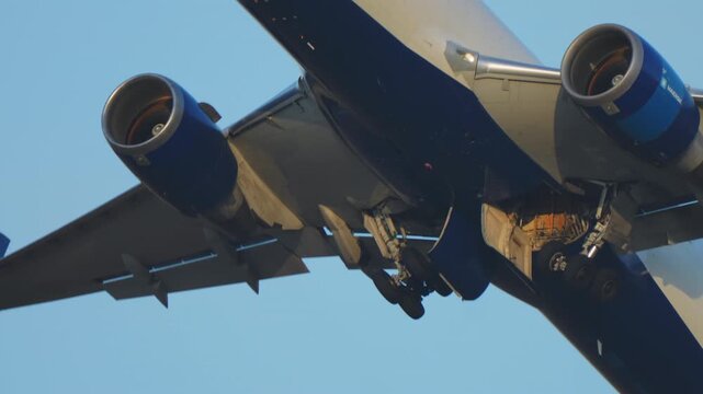 Extreme close-up of a wide-body commercial jet at the moment of takeoff with landing gear beginning to retract, twin engines with blue nacelles dramatically lit against a warm sky