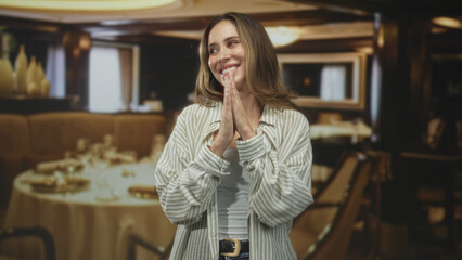 Woman smiling with hands clasped near chin over dining table in building wearing striped shirt and...