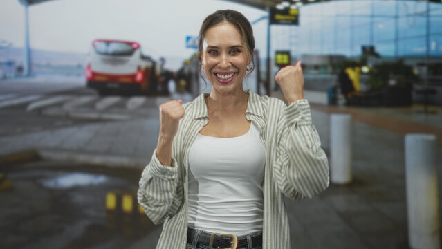 Woman smiling with thumbs up and raised fist, wearing white tank top at airport terminal; confidence travel.
