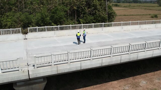 Concrete bridge construction inspection on empty roadway with two workers in safety vest and hard hat, rural landscape aerial view, infrastructure safety roadway bridge deck inspection