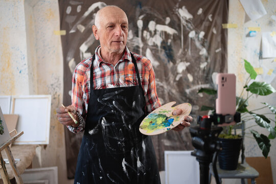 Senior elderly painter standing with drawing palette making video recording on smartphone using tripod in art creative studio.
