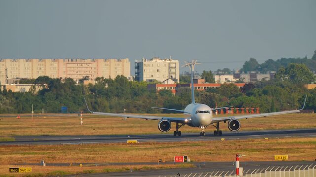 A wide-body twin-engine commercial jet taxis head-on toward the camera in warm golden hour light and starts accelerating, with runway signs and residential buildings bathed in orange glow in the