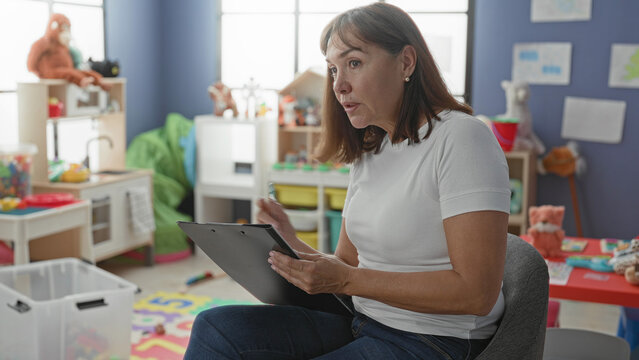 Woman writes on clipboard with pen amid colorful toys and cubbies in kindergarten building; focused observation.