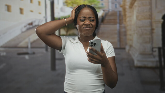 Woman holding smartphone, scratching head and grimacing while looking at the screen on a city street; digital frustration.