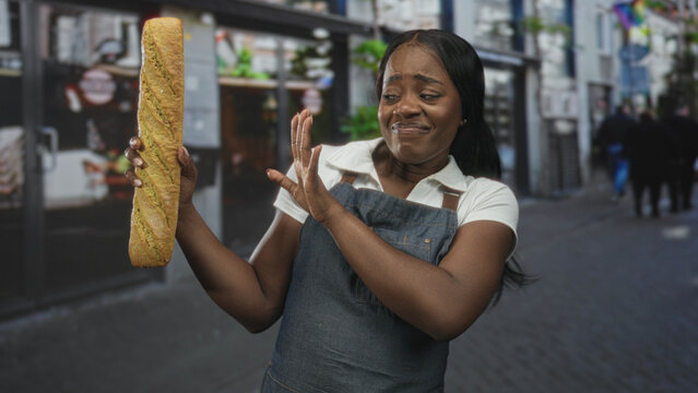 Woman baker in apron holds a baguette in her left hand and recoils with her right hand in a disgusted grimace on a street; disgust hesitation.