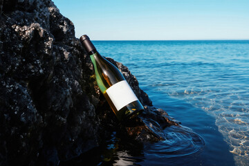 Obraz premium White wine bottle rests against dark wet rock at ocean edge. Clear blue water reflects sunlight. Clean label ready for branding text. Serene seascape.