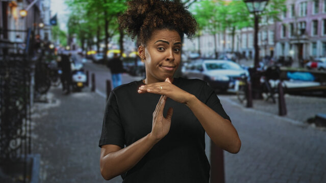 Woman with hands in t for timeout on city street, wearing black t shirt and looking skeptical while making a stop gesture; dismissal.