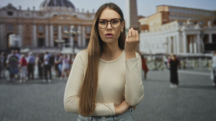Obraz premium Young woman with glasses holding up hand in pinched fingers gesture in front of building, crowded piazza and basilica visible; impatience tourism.