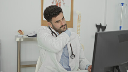 Fototapeta premium Man doctor rubbing his neck with stethoscope at clinic building desk using computer, indicating neck pain and discomfort; fatigue stress.