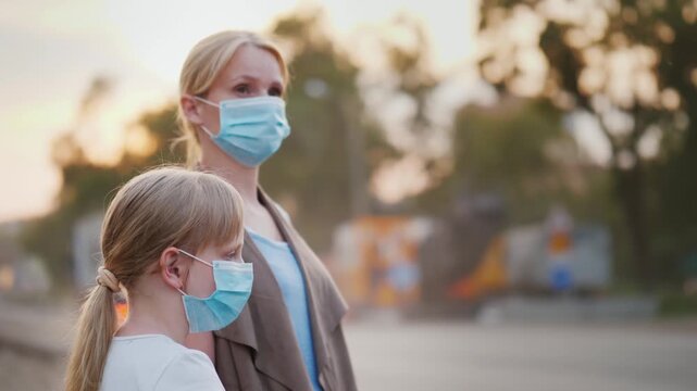 Mother and daughter in medical masks stand near busy road, cars passing by at golden hour, showcasing urban life during health crisis.