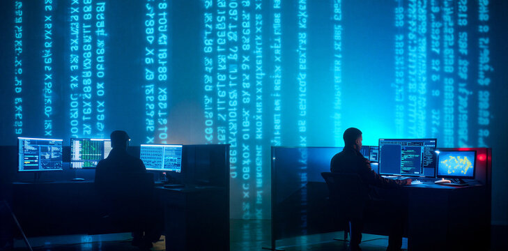 Cybersecurity experts working at computers in dark control room with large projected digital data streaming binary code on wall cybersecurity technology computer security