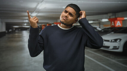 Naklejka premium Hispanic man points finger up and scratches head in an underground parking building beside parked cars and concrete pillars marked a, looking puzzled; confusion, searching.