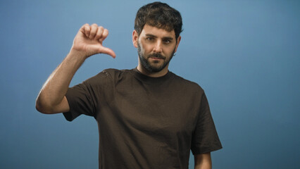 Fototapeta premium Hispanic young man with beard wearing brown t shirt showing thumb down with right hand in studio; disapproval critique.