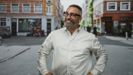 Fototapeta premium Man smiling and pointing hand toward camera on a city street wearing glasses and a white patterned shirt; confidence.