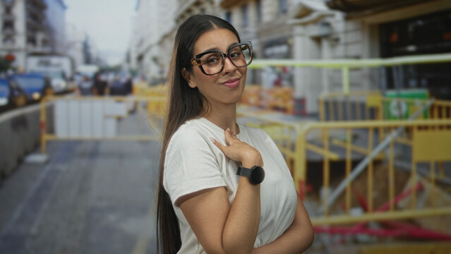 Woman in glasses wearing white t shirt and smartwatch, hand to chest and finger pointing to herself on street near construction barriers; self pride.