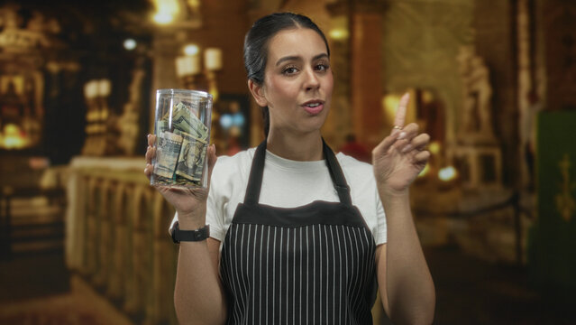 Woman in apron holding jar of money and pointing index finger in building; charity appeal compassion.