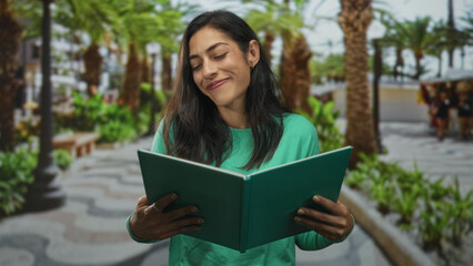 Fototapeta premium Woman reading green book and smiling while holding it on a sunny street lined with palm trees, wearing green sweater; contentment.