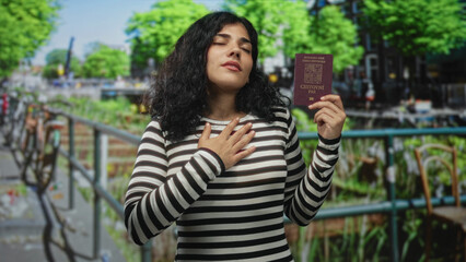 Fototapeta premium Woman holding czech passport on street bridge by canal, hand to chest with eyes closed and relaxed face; pride identity.