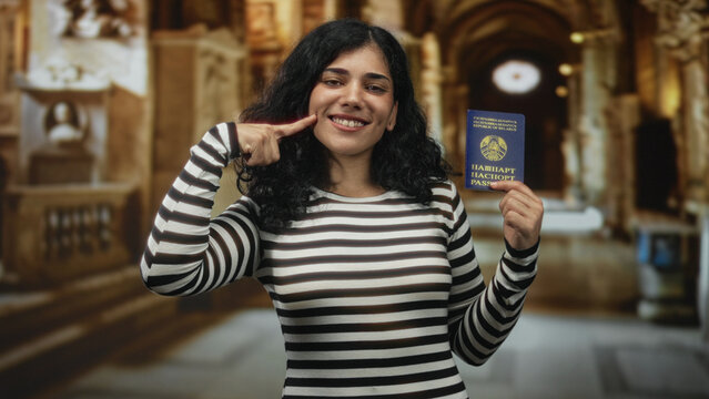 Woman smiling, holding belarus passport and pointing finger to cheek in church building interior; identity travel pride.