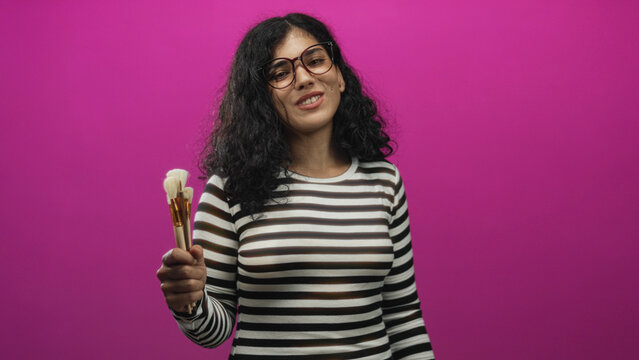 Young arab brunette woman wearing glasses holding makeup brushes in one hand against a pink backdrop in studio, smiling and posing; playful beauty creativity.