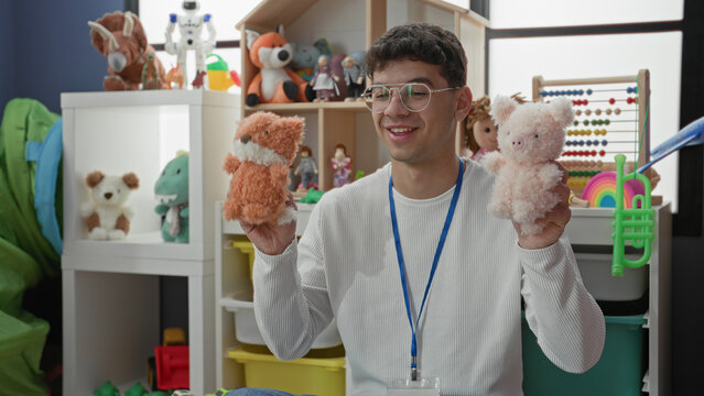 Man teacher wearing lanyard and glasses holding two plush hand puppets amid toys and abacus in a playroom inside a building; playful learning.